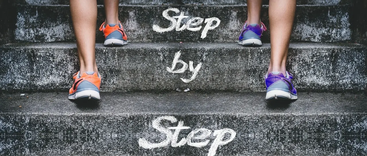 Nohy v teniskách stoupají po betonových schodech s nápisem „Step by step“